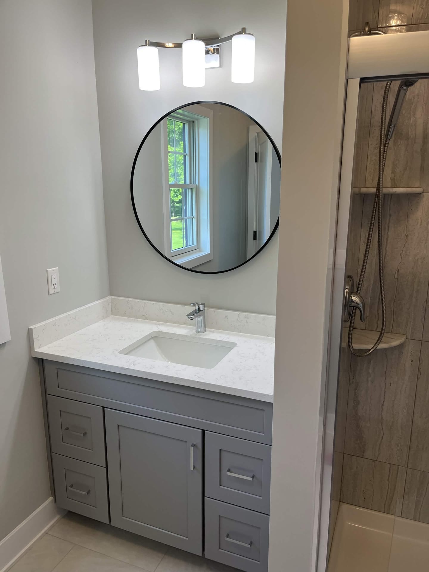 Modern bathroom vanity with round mirror, wall lighting, gray cabinetry, quartz-style countertop, and tiled shower, built by Cursi Construction Inc.