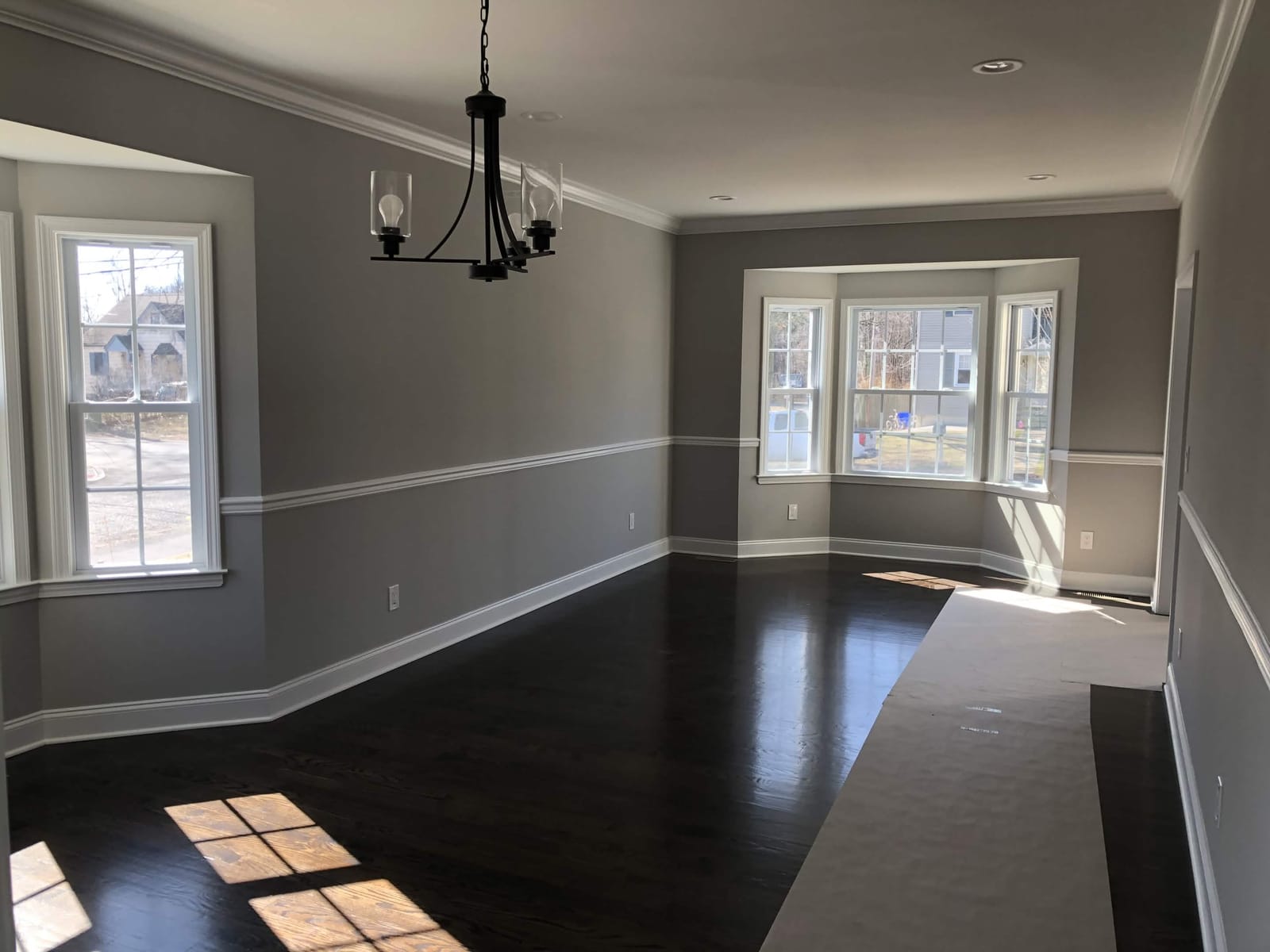 Finished dining room with gray walls, white crown molding and chair rail, dark hardwood floors, large windows, and black chandelier, built by Cursi Construction Inc.