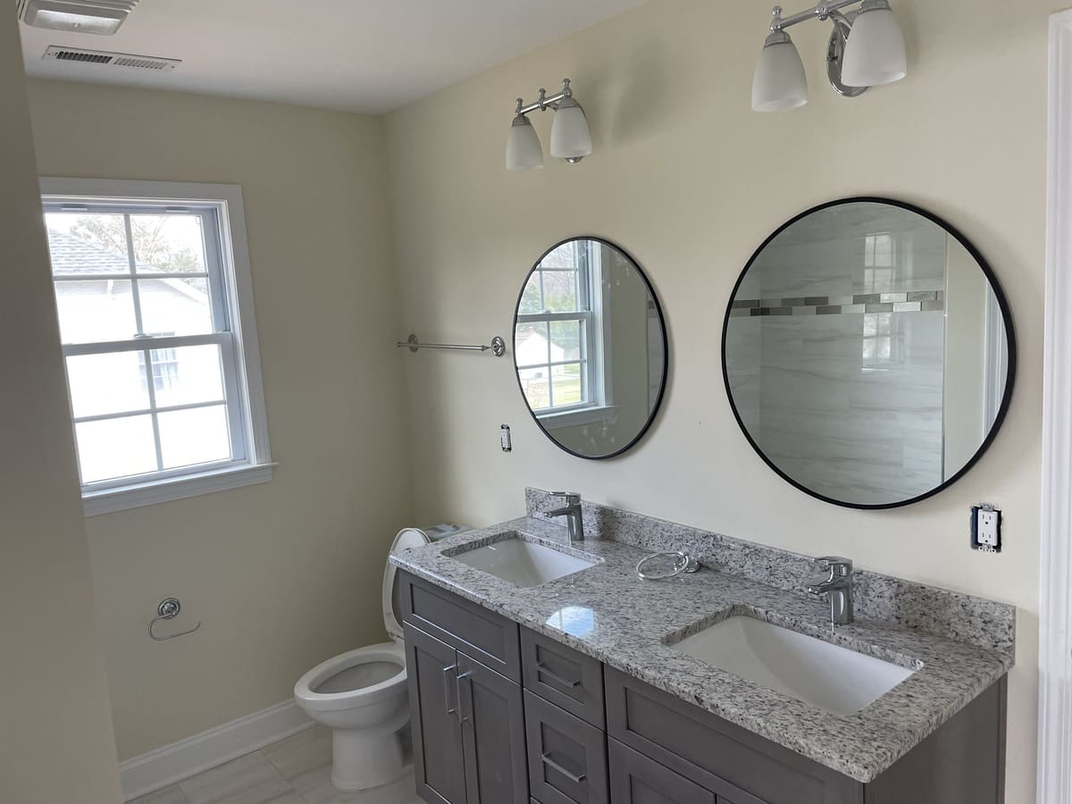 Bathroom renovation with double gray vanity, granite countertop, two round mirrors, and wall lights, built by Cursi Construction Inc.