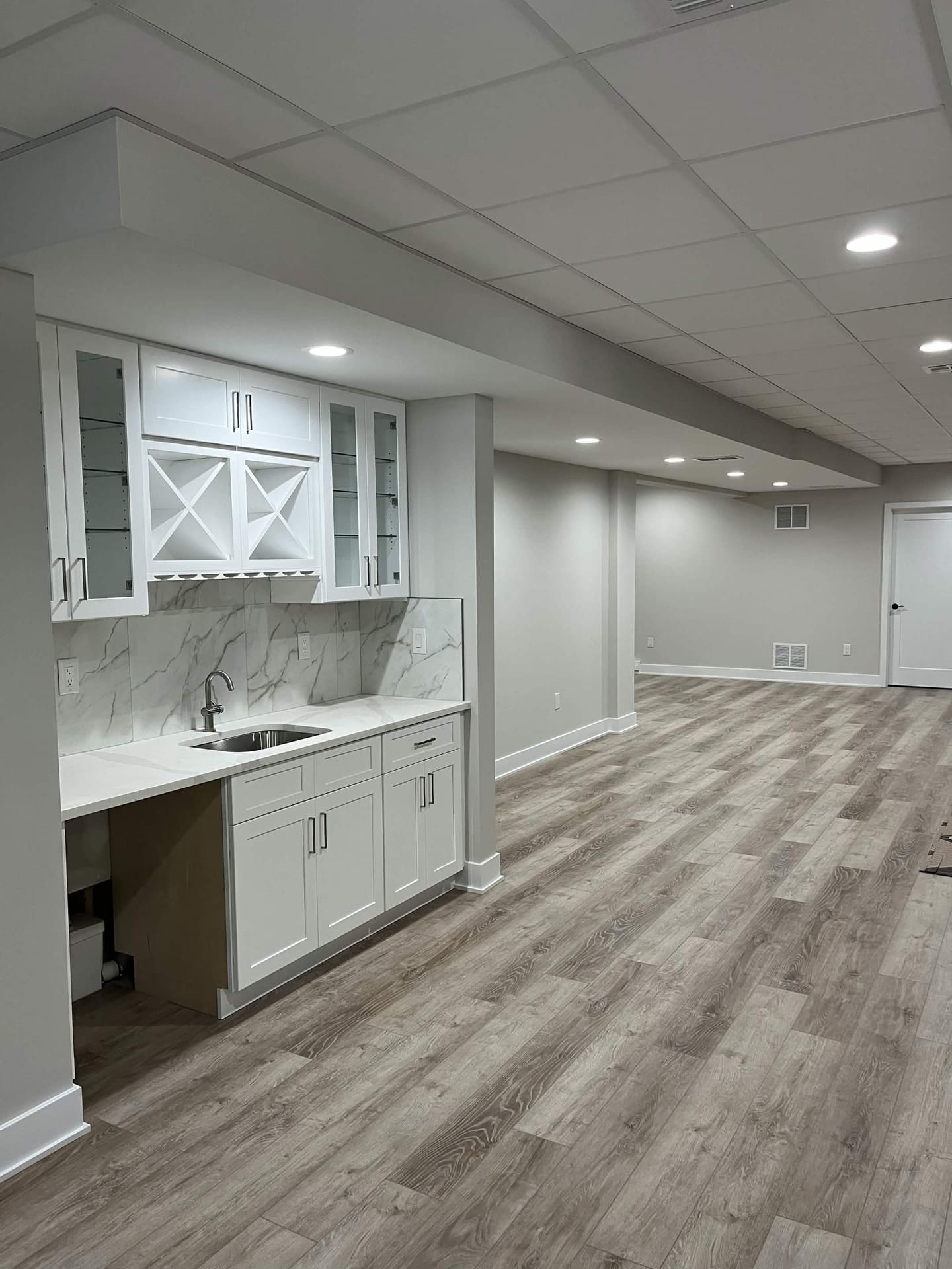 Finished basement wet bar with white cabinetry, marble-look backsplash, and LVP flooring by Cursi Construction Inc.